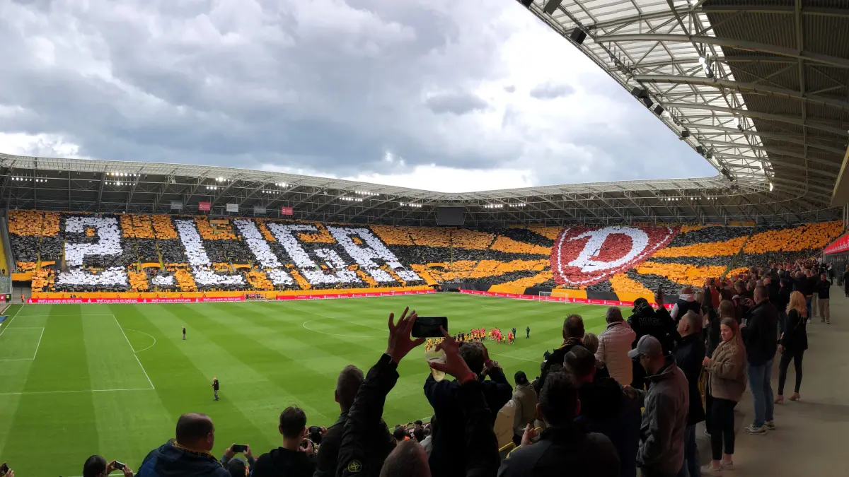 Fußballfans stehen in einem Stadion, halten Fanartikel, die ein Mosaik bilden.