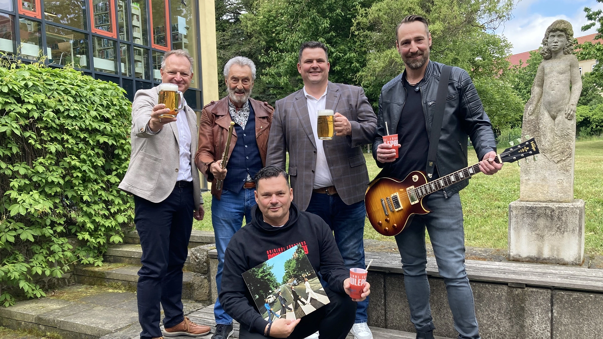 Fünf Männer stehen lächelnd im Freien, einer hält eine Gitarre.