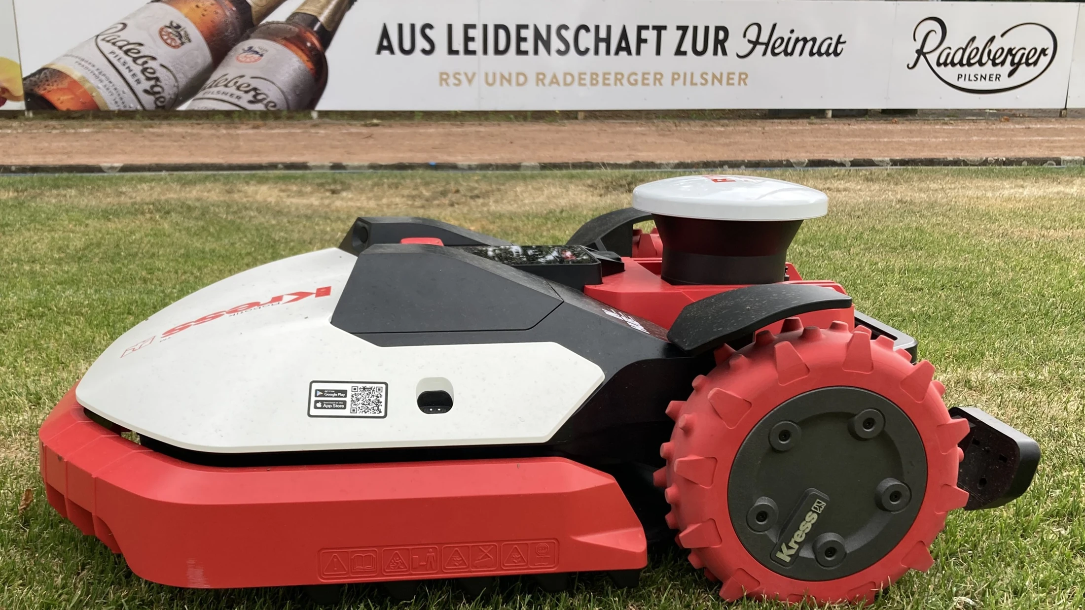Rasenroboter steht auf einer gemähten Rasenfläche. Im Hintergrund eine Werbebande von Radeberger.
