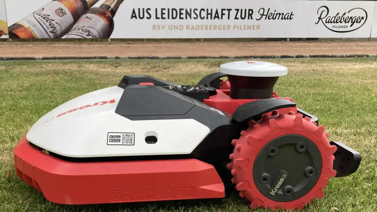 Rasenroboter steht auf einer gemähten Rasenfläche. Im Hintergrund eine Werbebande von Radeberger.