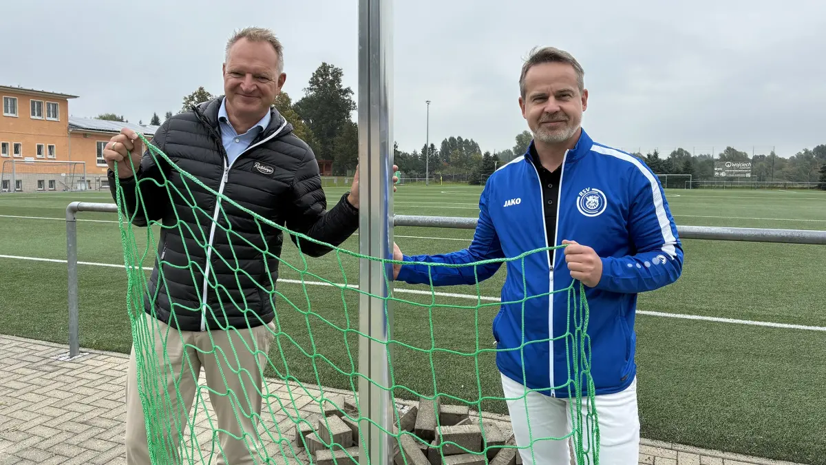 Zwei Männer halten ein grünes Netz am Rand eines Fußballplatzes. Im Hintergrund liegt das Spielfeld.