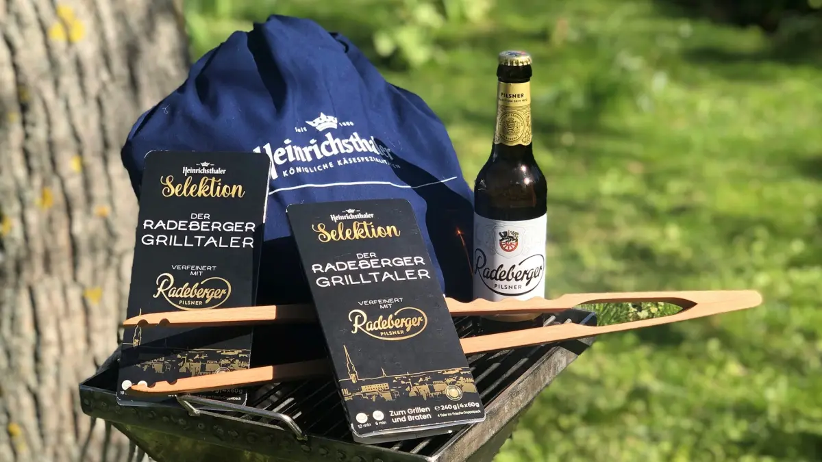 Grillzubehör mit Radeberger Grilltaler-Verpackung und Radeberger Pilsner-Flasche auf einem Grill im Freien bei Sonnenschein.