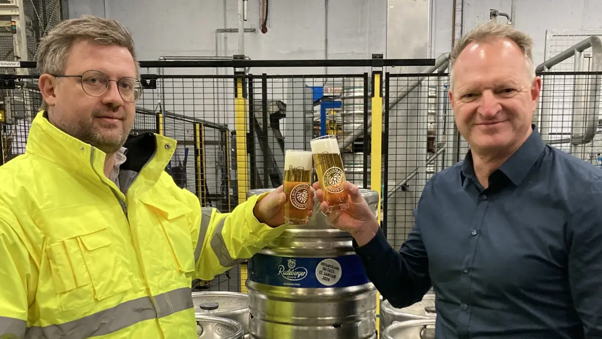 Zwei Personen in einer Brauerei, die Biergläser mit Schaum anstoßen. Einer trägt eine gelbe Jacke, der andere ein dunkles Hemd.