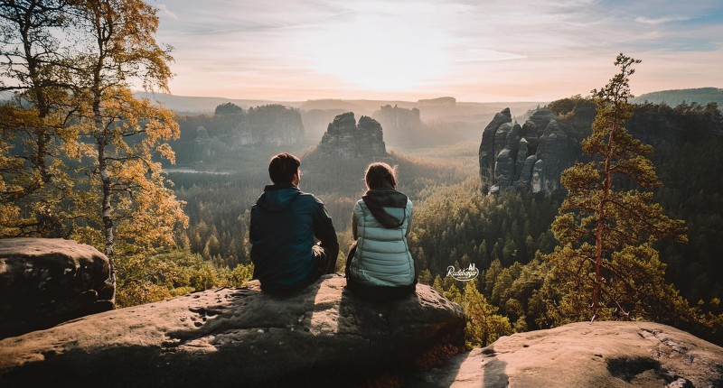 Zwei Personen sitzen auf einem Felsen und blicken auf eine Felslandschaft im Nebel.