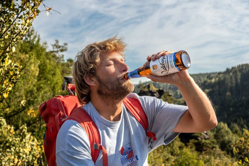 Ein Wanderer mit Rucksack trinkt aus einer Flasche vor einer bewaldeten Hügellandschaft.